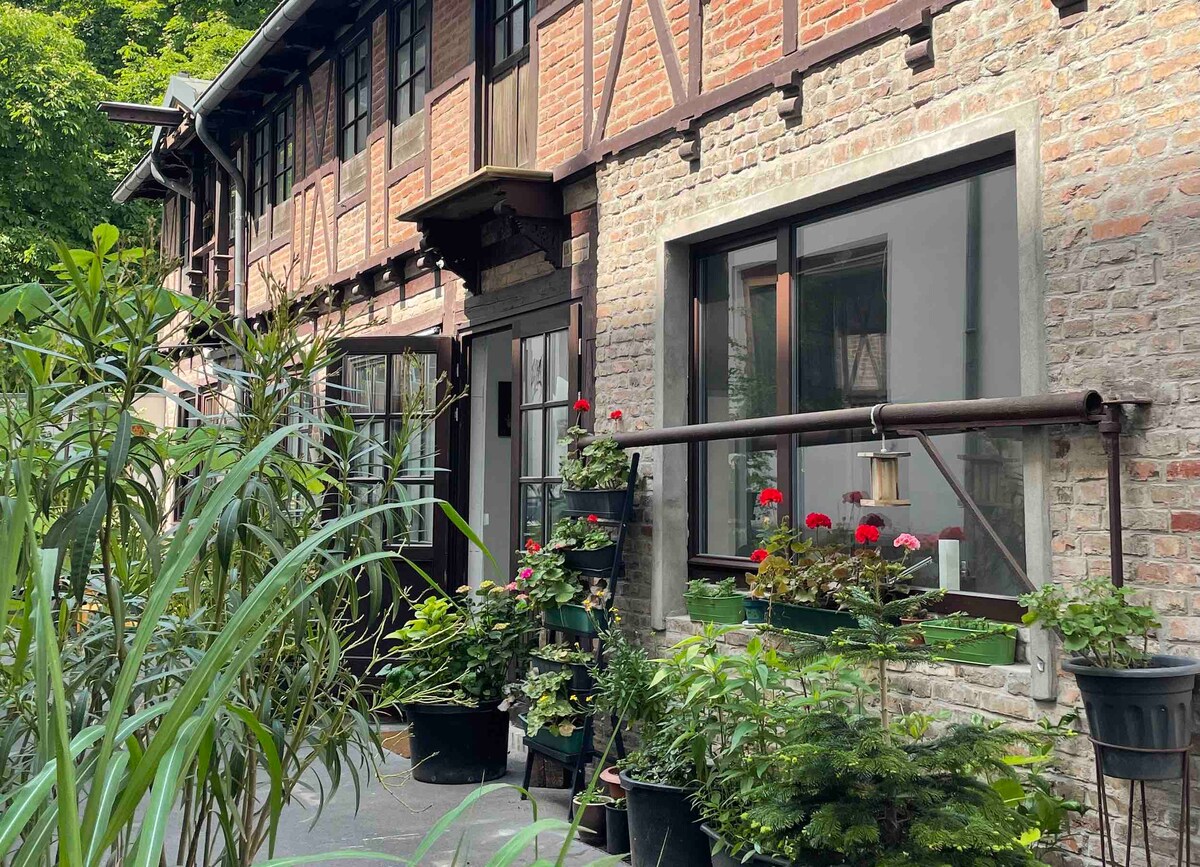 The exterior of the renovated building features exposed brick walls and wooden beams, surrounded by greenery. Numerous potted plants with vibrant flowers create a welcoming entryway, while large windows provide glimpses into the interior. The setting is serene, situated in a quiet courtyard.