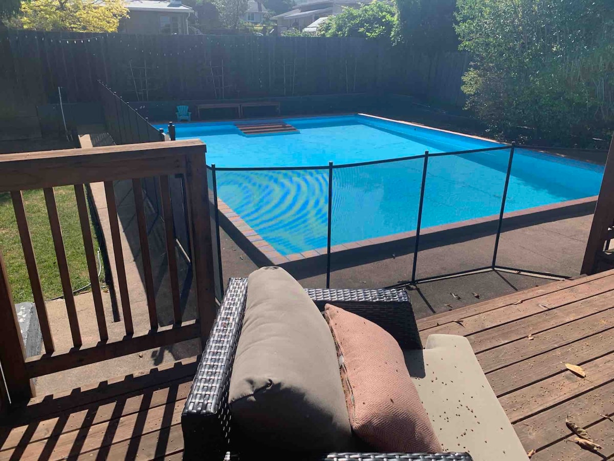 A clear blue fenced-in pool is visible from a wooden deck area. A comfortable seating option with cushions is positioned nearby. Lush greenery surrounds the pool area, adding to the outdoor setting. A sunlit patio space is also seen in the foreground.