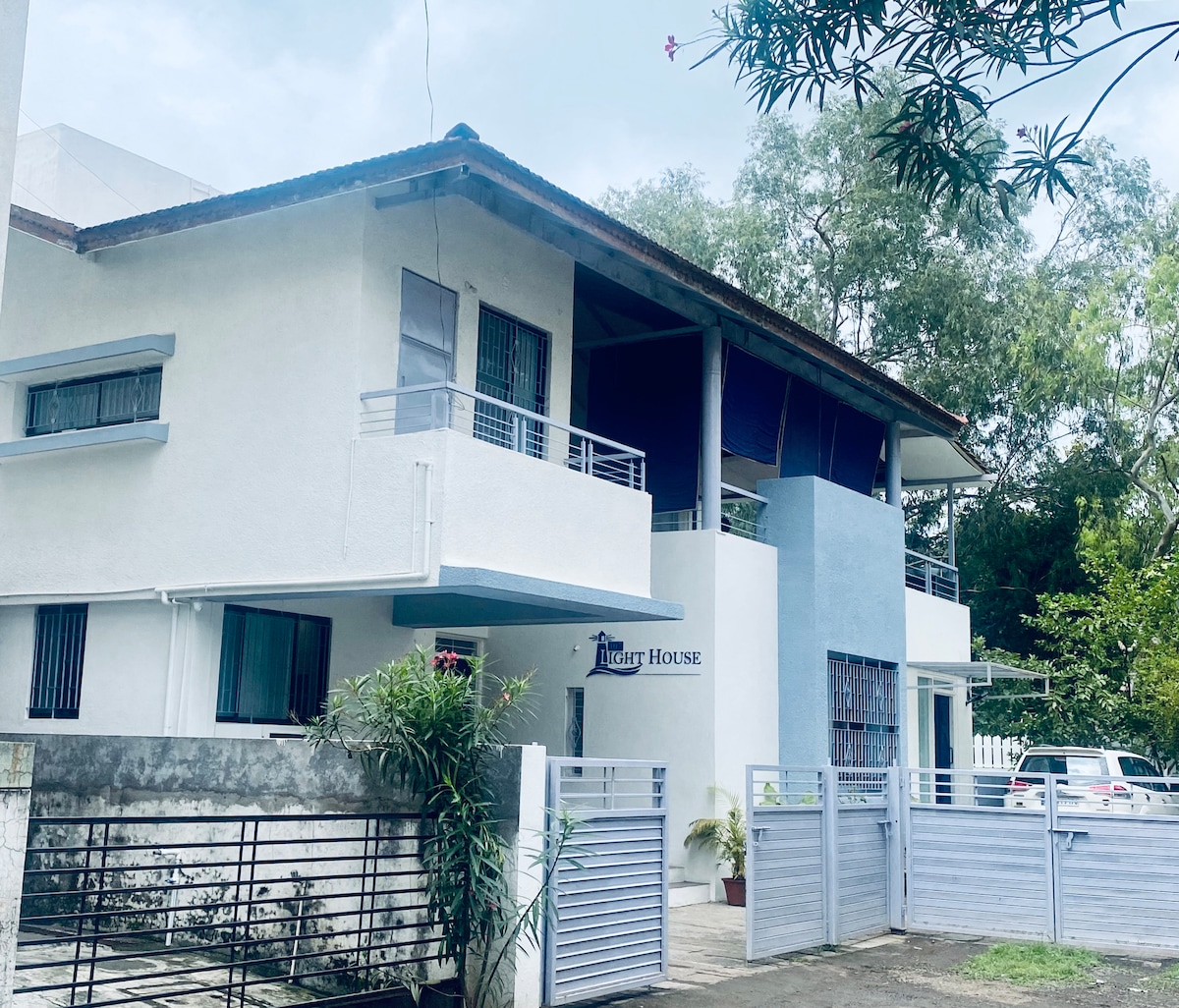 The exterior of the villa features a two-story structure with a light-colored façade. A covered balcony is visible on the upper floor, and a parking area with space for several vehicles is accessible in front. Lush greenery surrounds the property, enhancing the serene ambiance.