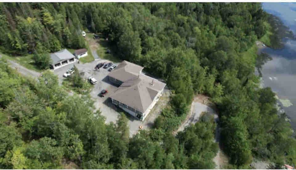 An aerial view of the cottage property reveals a spacious building surrounded by lush green trees. A gravel driveway leads to the entrance, with several parked cars nearby. The serene lake can be seen at the edge of the property, emphasizing the natural setting.