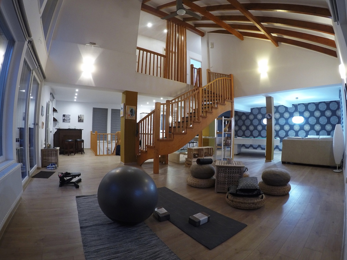 An open living area features a wooden staircase leading to an upper level. Cozy seating is provided by various round poufs and a large exercise ball. Natural light fills the space through large glass doors, highlighting the modern decor and spacious layout.