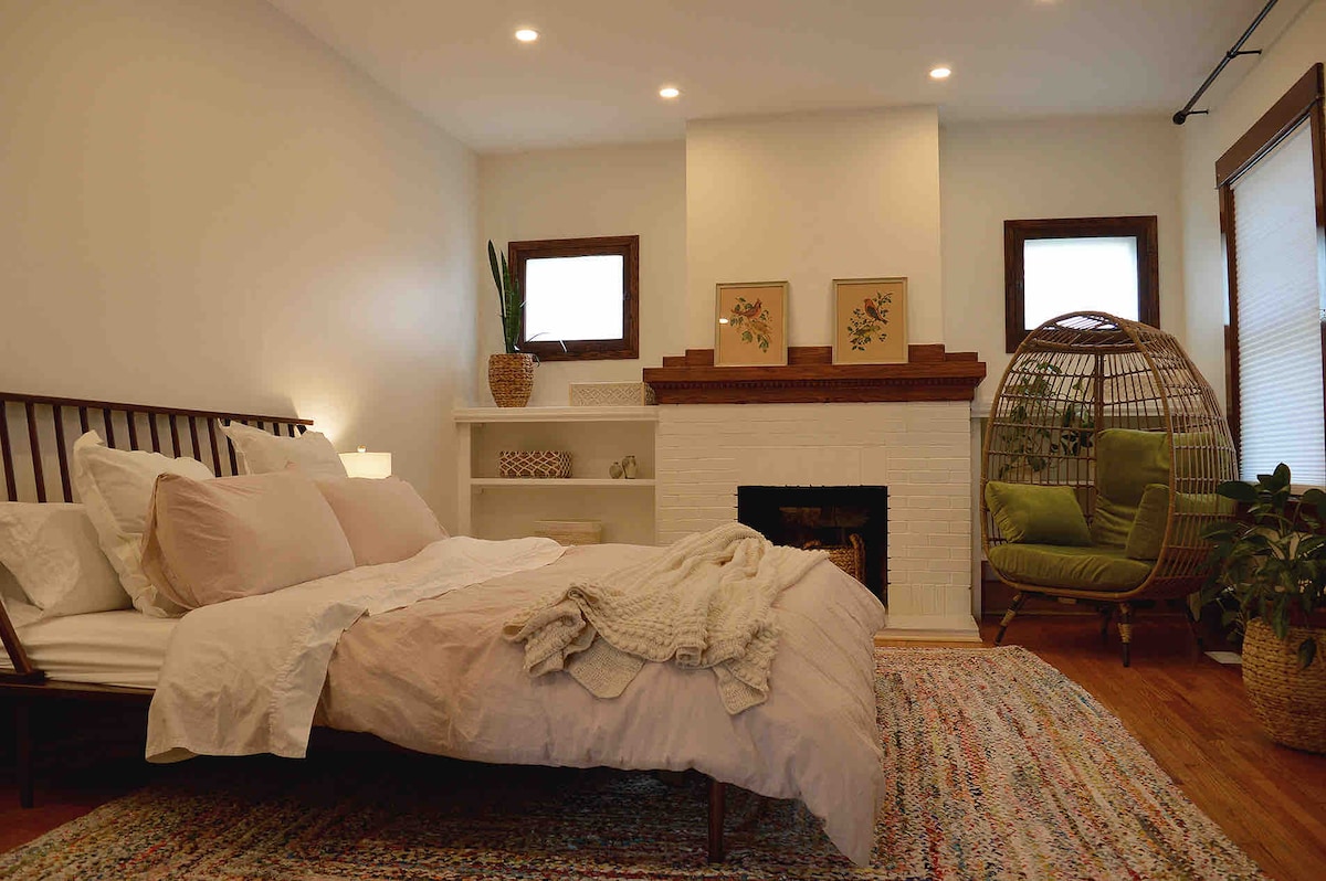 A spacious bedroom features a king-size bed with soft linens, placed alongside a decorative fireplace. Natural light enters through two windows, and a green armchair accentuates the space. A colorful area rug and potted plants add warmth and texture to the inviting atmosphere.