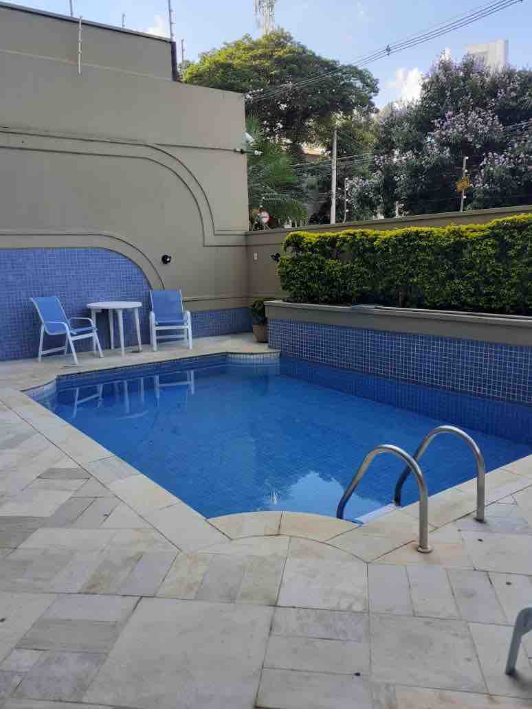 A small swimming pool with a clear blue surface is surrounded by neatly arranged stone tiles. A set of two blue chairs and a white table is positioned nearby, offering a space for relaxation. Lush greenery and landscaped hedges provide a natural backdrop.