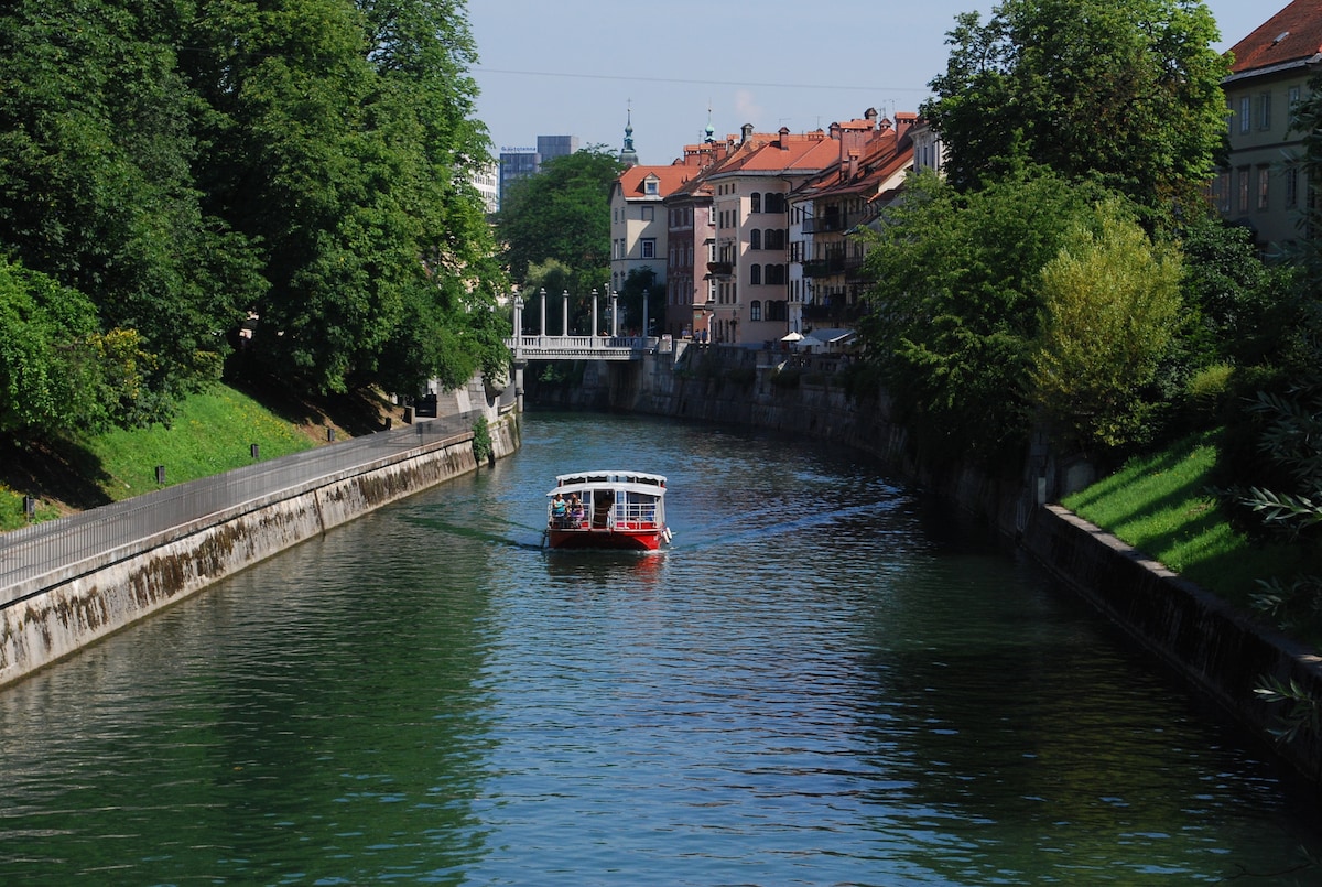 A boat moves gently along the Ljubljanica River, surrounded by lush greenery and historical buildings. The water reflects the blue sky and vibrant colors of the surrounding structures, while pedestrians can be seen enjoying the riverbank path.
