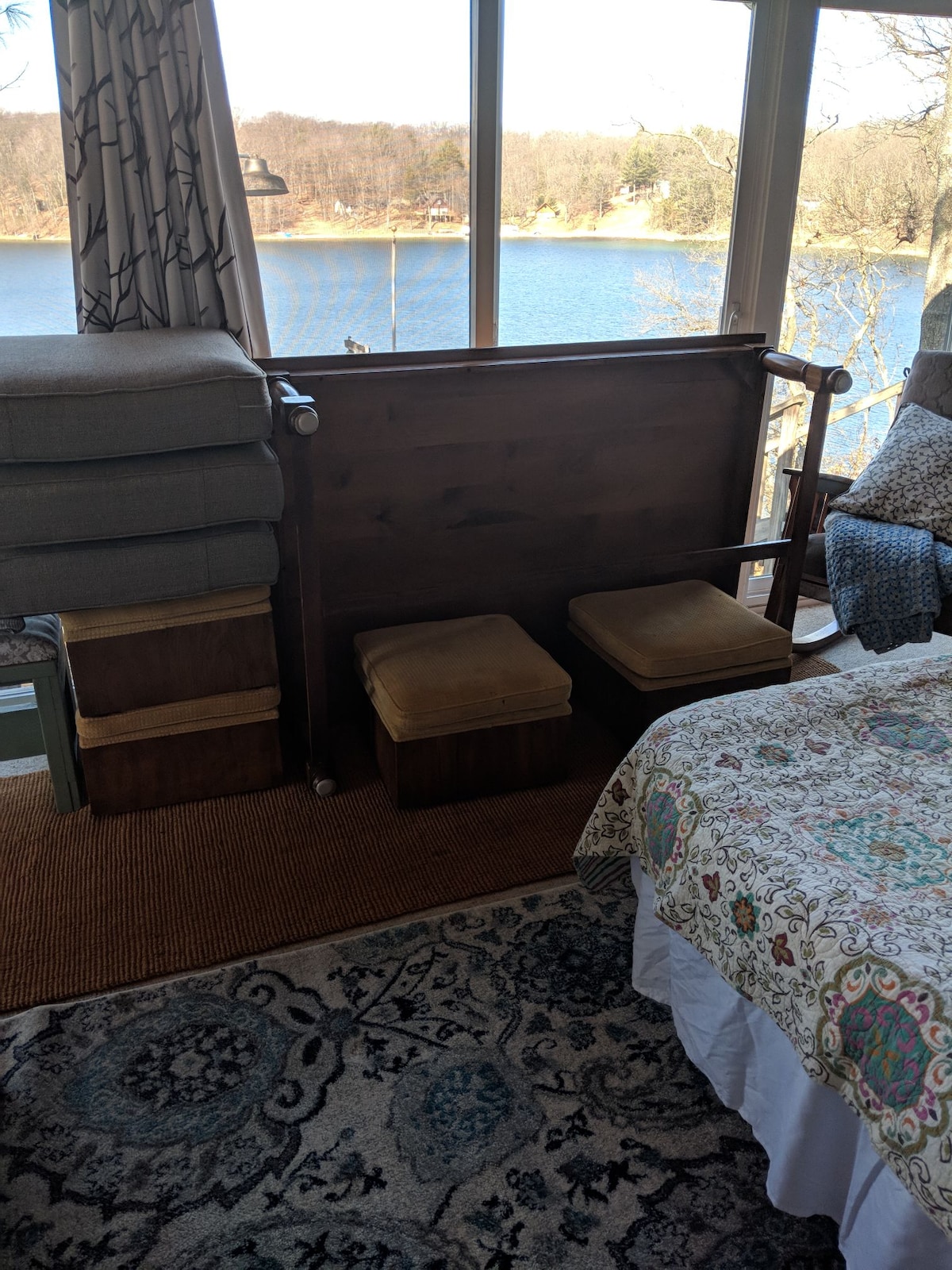 A cozy sleeping area features a spacious bed adorned with a patterned quilt. In the background, lake views are framed by large windows. Additional seating is provided by two small stools and a side table, contributing to the inviting ambiance of the space.