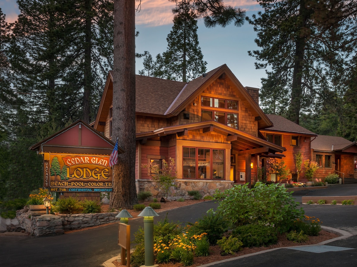 The exterior of Cedar Glen Lodge is showcased, featuring a rustic wooden design accentuated by warm lighting. Large trees are visible around the property, and a welcoming sign identifies the lodge. A paved path leads towards the entrance, framed by natural landscaping.