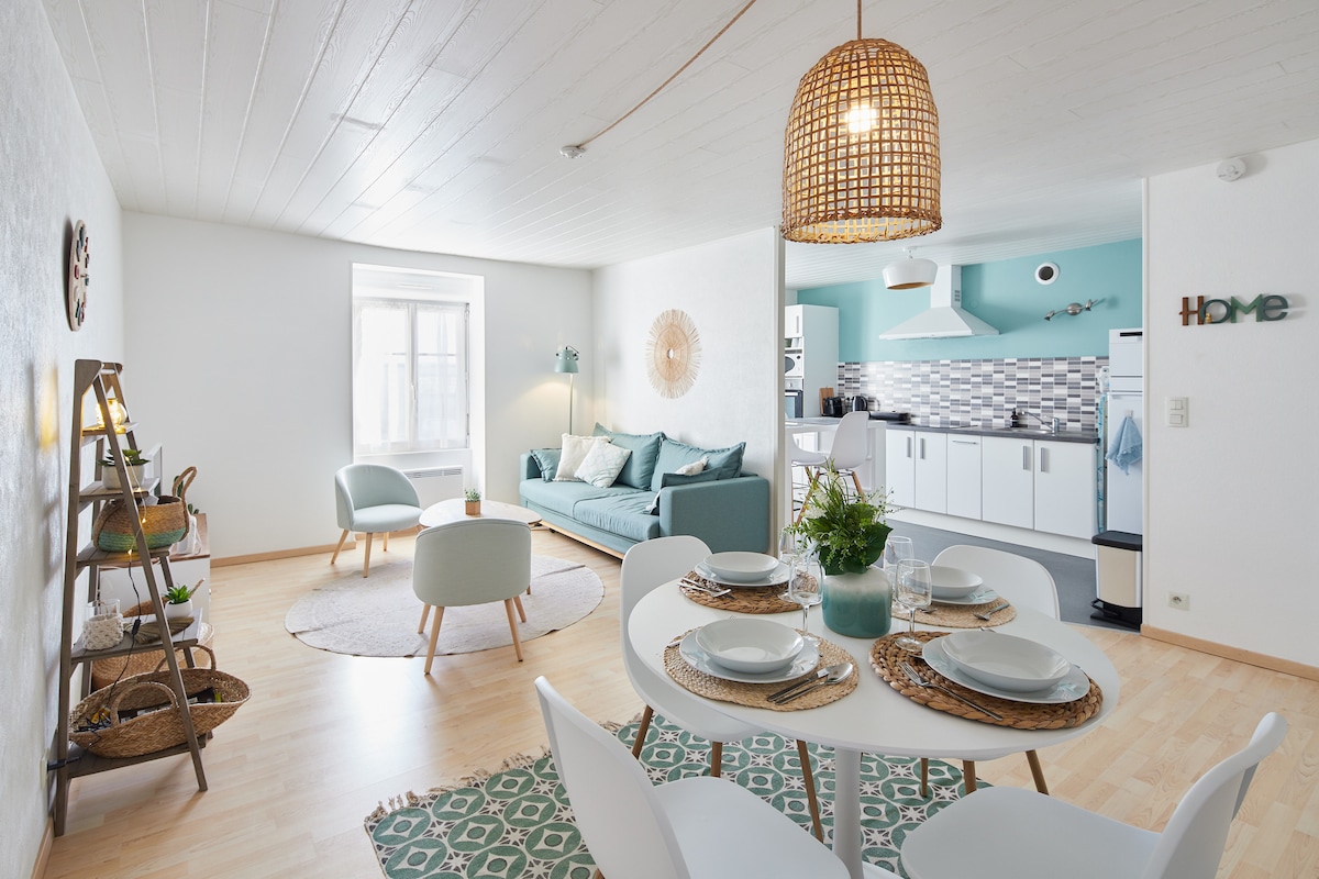A bright living space featuring a soft green sofa with decorative cushions, a round rug beneath, and a dining table set for four. The open kitchen is visible in the background, with modern appliances and a light color scheme that enhances the minimalist Scandinavian design.