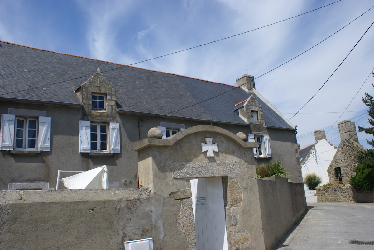 A charming stone house is depicted with a grey façade, framed by light blue shutters. A decorative stone archway leads to the entrance, and a path runs alongside the structure. The sky is partly cloudy, providing a serene backdrop to the home.