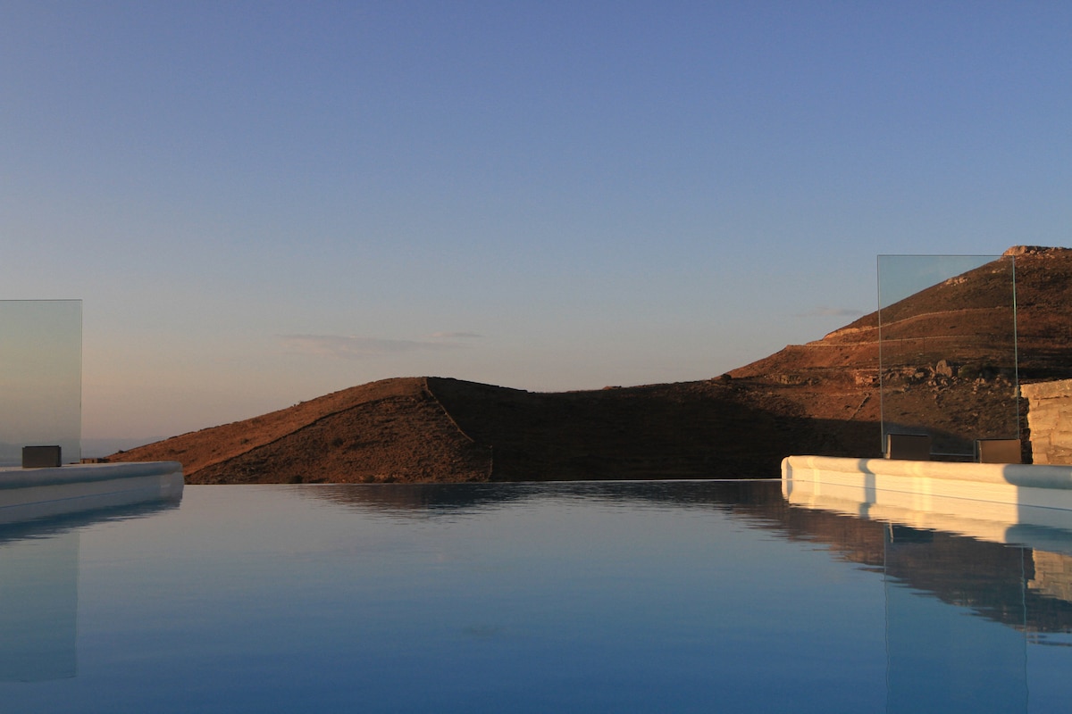 An expansive view of the tranquil outdoor swimming pool is captured, reflecting the soft colors of the evening sky. Surrounding hills are visible in the background, adding a serene natural landscape to the setting.