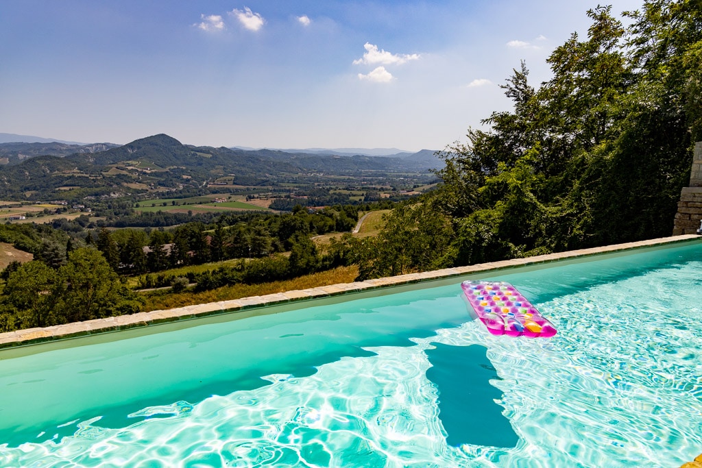 The image shows a glistening saltwater pool, reflecting sunlight with a colorful floating mattress on the surface. Surrounding hills and valleys create a picturesque backdrop, filled with lush greenery and distant mountains under a clear blue sky.