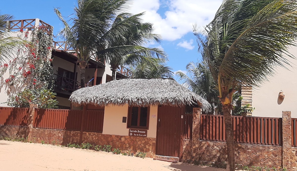 A charming structure features a thatched roof and wooden walls, surrounded by tropical vegetation. Lush palm trees sway gently in the breeze, while a wooden fence offers privacy. The bright blue sky enhances the welcoming atmosphere of this outdoor space.