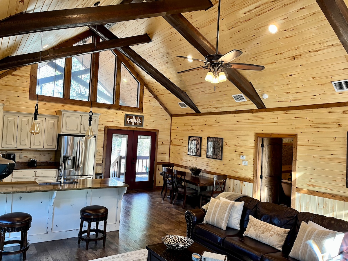 The open-concept living area features vaulted wooden ceilings, exposed beams, and large windows that invite natural light. A well-equipped kitchen with modern appliances can be seen alongside a cozy seating area, which includes a leather sofa and a dining table set for four.