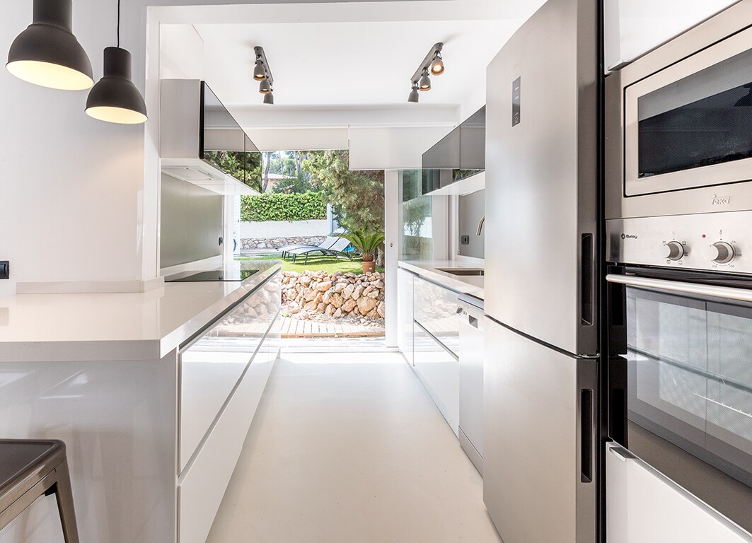 The modern kitchen is equipped with sleek, white cabinetry and stainless steel appliances. Large windows provide a view of the outdoor area, while the space is well-lit by pendant lighting. An organized arrangement enhances functionality, making it a welcoming environment for meal preparation.