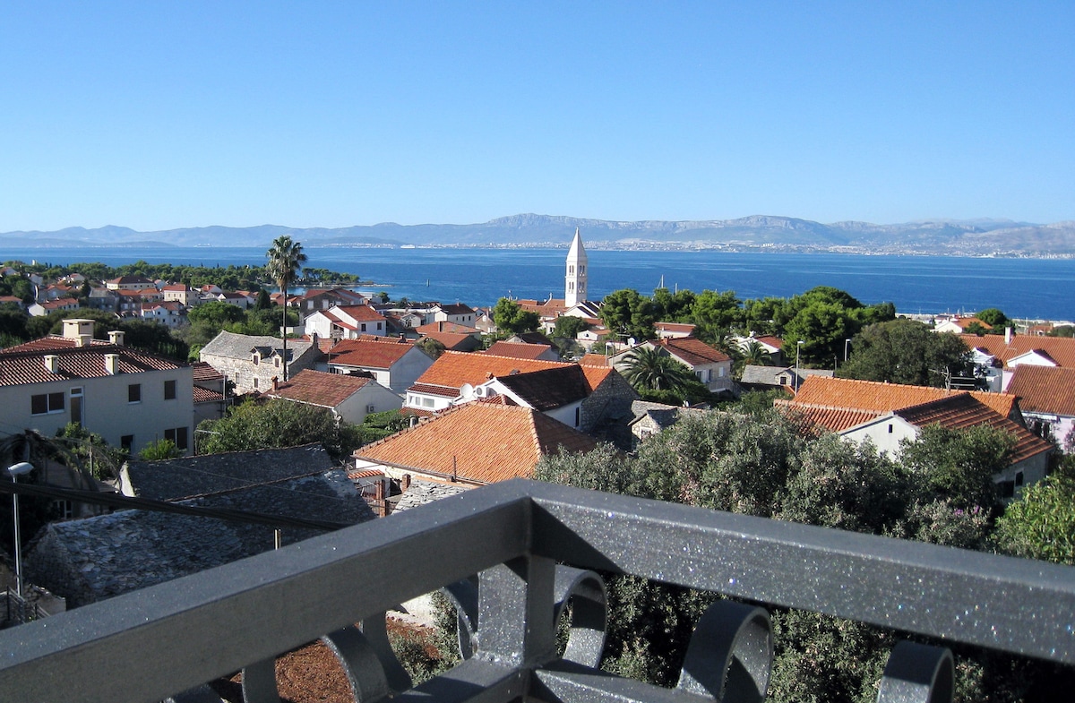 A view from a private balcony reveals a scenic landscape of terracotta-roofed houses and lush greenery. In the background, a distant church spire stands out against the blue sky, while the sea glistens under sunlight. The horizon is defined by gentle mountain silhouettes.