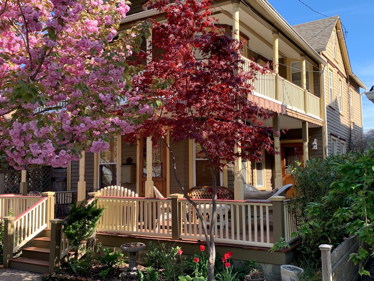 A charming Victorian home adorned with blooming pink cherry blossoms and vibrant red foliage. The wrap-around porch features seating arrangements and is framed by lush greenery, creating a warm and inviting entrance. The exterior showcases a blend of historic architecture and rejuvenated elements.