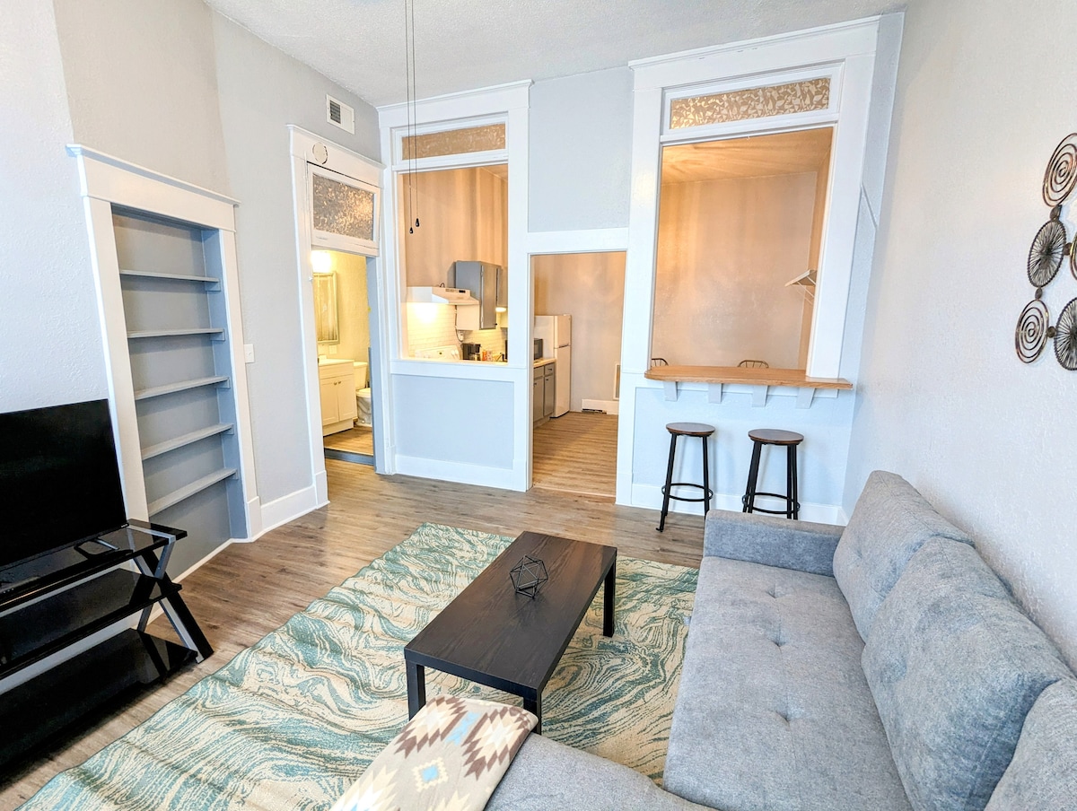 A comfortable living space features a gray sofa and a black coffee table, complemented by a patterned area rug. A breakfast bar with two stools is visible, along with an open view into the kitchen area, while large windows allow natural light to fill the room.