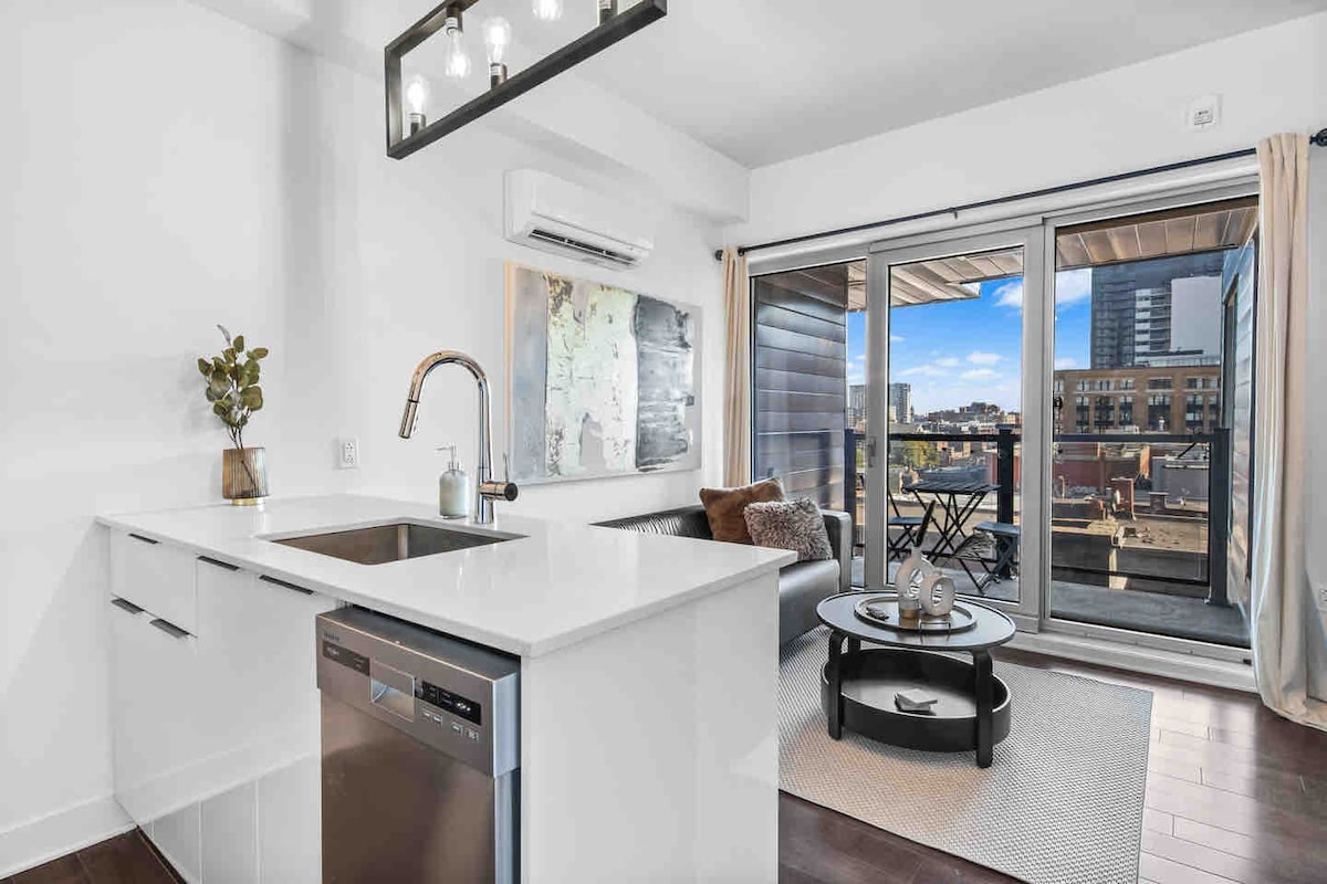 A modern kitchen area features sleek white cabinetry and a stainless steel dishwasher. Natural light floods the space through glass sliding doors that open to a balcony with outdoor seating and city views. A textured area rug adds warmth to the floor.