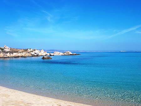 A tranquil seascape showcases calm blue waters meeting a sandy beach. The shoreline is lined with smooth stones, while gentle waves create a serene atmosphere. A clear sky with hints of distant land adds to the peaceful ambiance of the coastal scene.