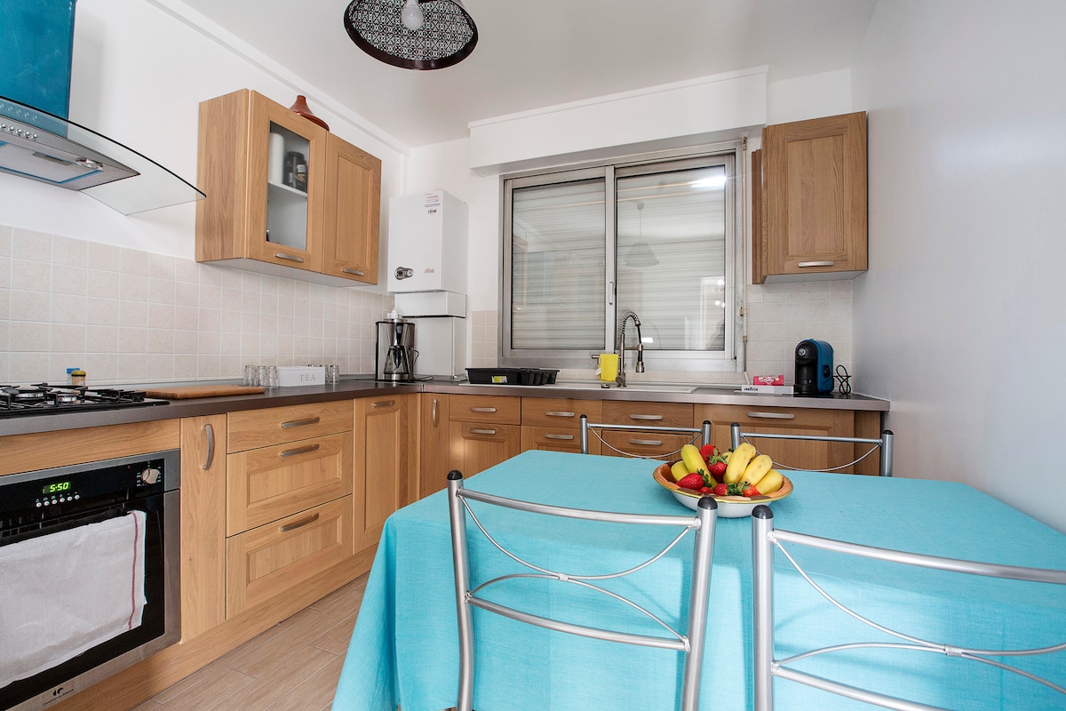 A modern kitchen features warm wooden cabinetry and a spacious countertop. A round table is set with a bowl of assorted fresh fruit. A window offers natural light, and appliances such as a coffee maker and kettle are visible. The cooking area includes a gas stove and oven.