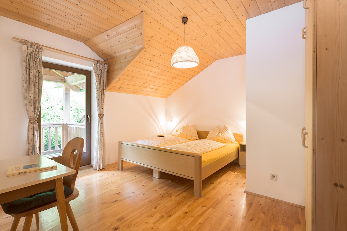 A cozy bedroom features a wooden ceiling and warm wood flooring. Two spacious beds are adorned with yellow bedding. A small wooden table is positioned near a window that opens to a private balcony, allowing natural light to fill the room.
