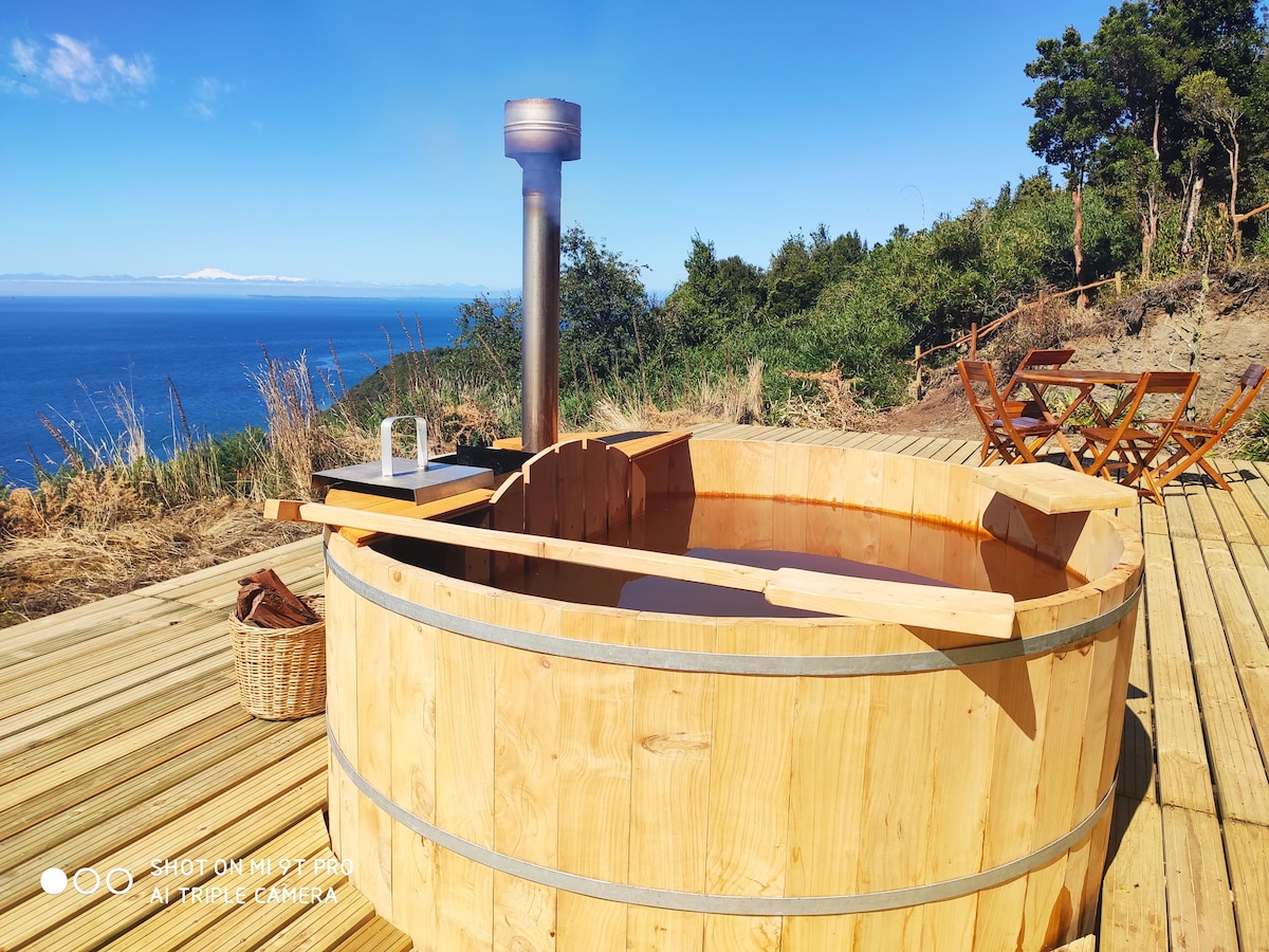 A wooden hot tub is positioned on a wooden deck, overlooking a scenic view of the ocean and distant mountains. A small table with chairs is visible nearby, enhancing the outdoor experience. The clear sky and lush greenery surround the area.