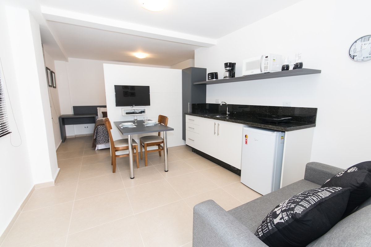 The studio features an open layout with a kitchenette equipped with essential appliances, including a refrigerator and microwave. A dining table with chairs is positioned adjacent to a comfortable sofa. A wall-mounted TV is visible, enhancing the living space's functionality.