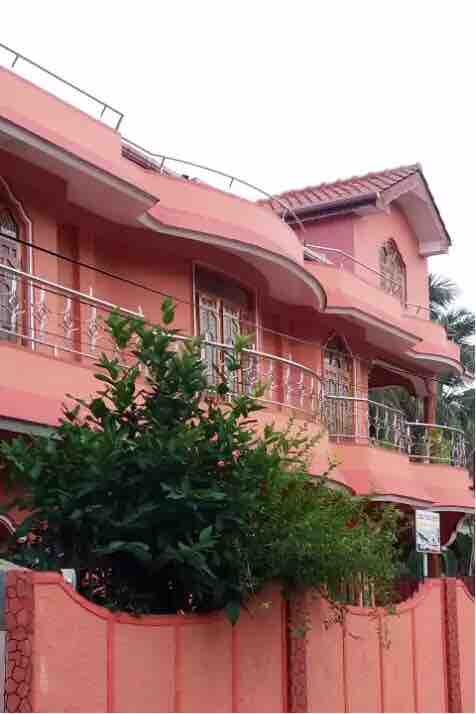 A two-story pink property features decorative balconies and arched windows, surrounded by lush greenery. A low fence frames the front entrance, enhancing the welcoming appearance of the home. A tiled roof adds to the architectural charm.