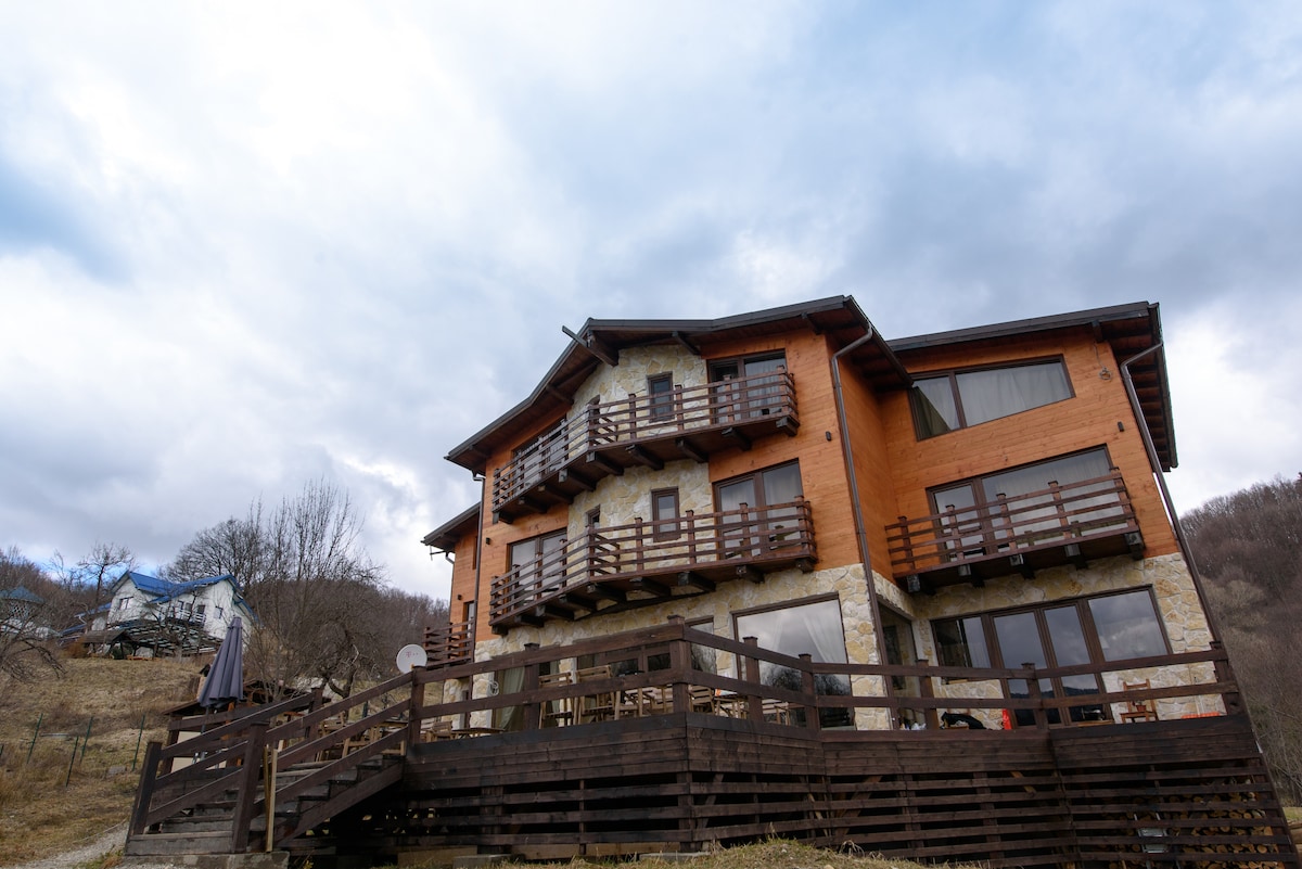 A charming multi-level house showcases a combination of wooden and stone elements. Several balconies with wooden railings extend from the upper floors. A spacious deck surrounds the lower level, providing an outdoor area with a view of the forested surroundings.
