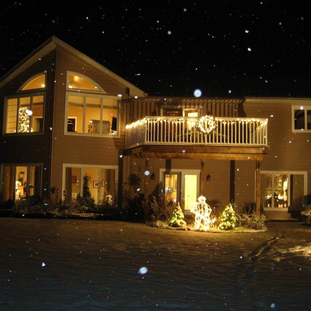 The exterior of the home is beautifully lit against a snowy backdrop. The balcony is adorned with string lights and festive decorations. Large windows reveal a warm glow from inside, complemented by snow-covered landscaping and a peaceful winter atmosphere.