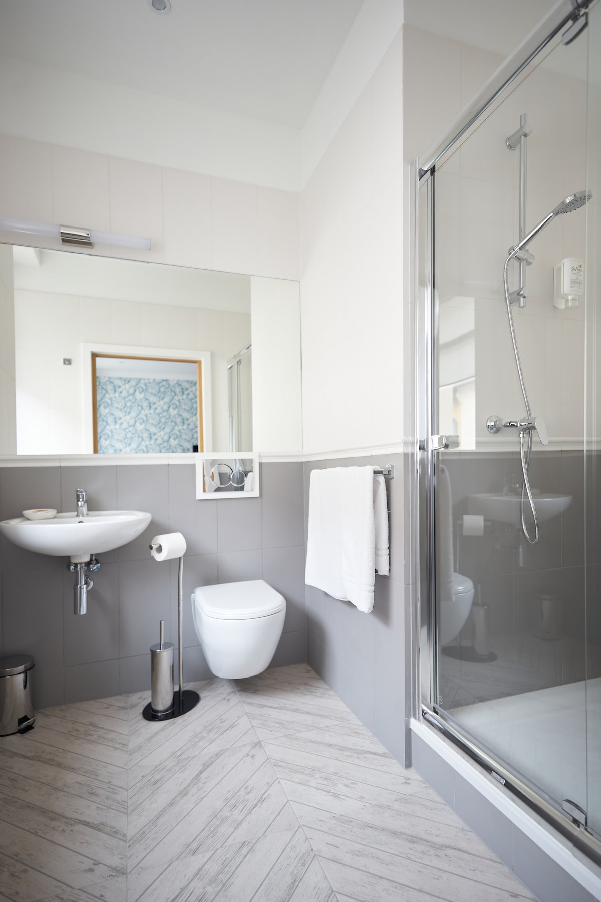 The bathroom features a walk-in shower, a modern toilet, and a sink with a large mirror above. Soft towels hang neatly by the shower. A waste bin and a toilet paper holder are included, with textured, light-colored floor tiles creating a clean and contemporary look.