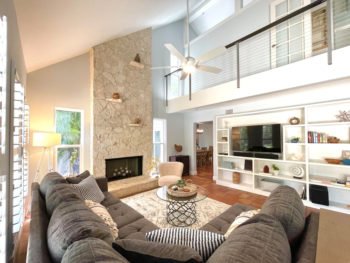 The living room features a large sectional sofa centered around a round coffee table. A stone fireplace contributes to the room's warmth, while tall windows allow natural light to fill the space. A ceiling fan provides airflow above the seating area.