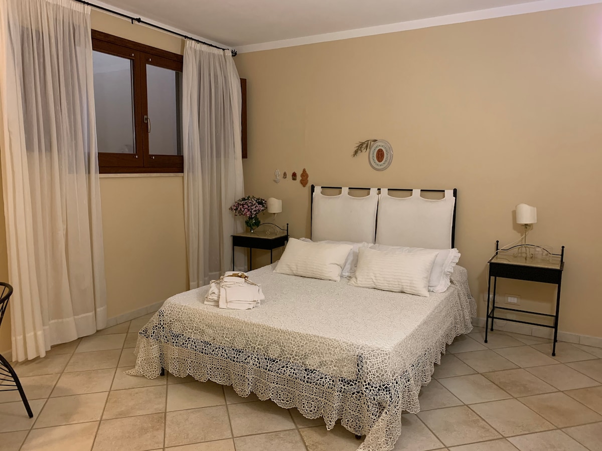 A spacious sleeping area features a large bed adorned with a light-colored knitted throw. Two bedside tables provide ambient lighting, while soft curtains frame the window, allowing natural light to enter. Neutral walls and tiled flooring contribute to a warm, inviting environment.