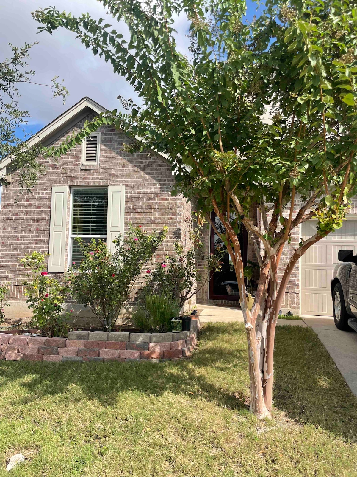A charming brick home is set amidst a well-maintained front yard featuring green grass and flower beds. Sunlight filters through the leaves of a young tree, while neatly trimmed bushes add to the inviting exterior. A driveway leads up to a garage on the right.