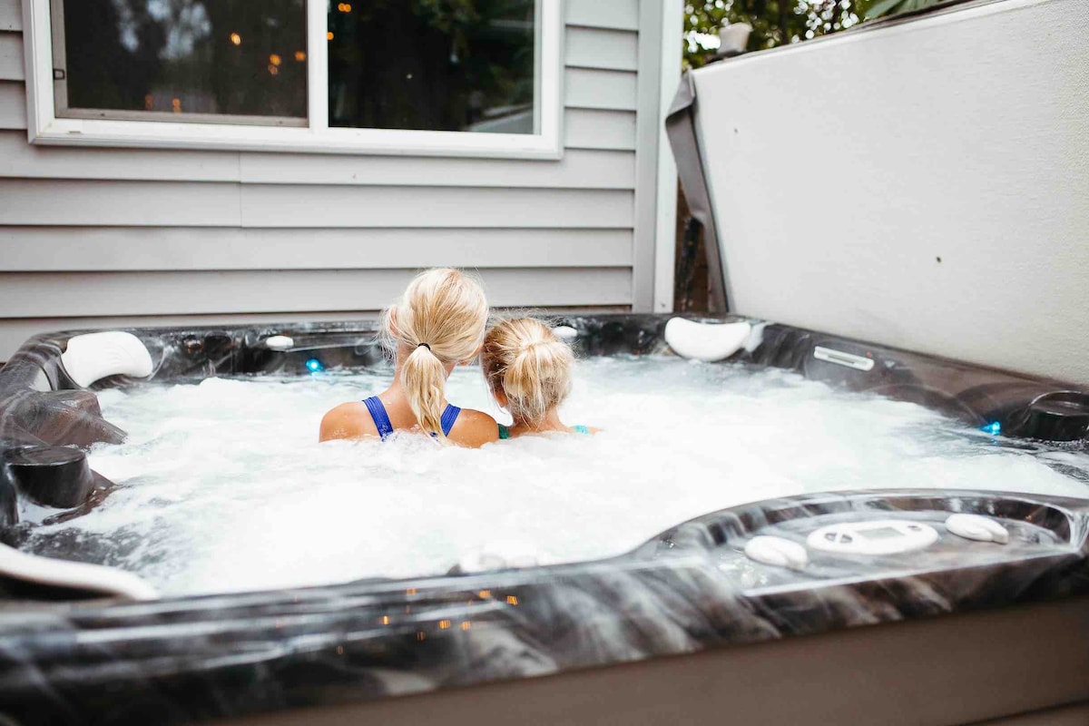 A private hot tub is nestled in a secluded outdoor area, surrounded by a gray exterior wall. Bubbles rise from the water, creating a inviting atmosphere. Two individuals are seated with their backs facing the camera, enjoying the tranquil experience.