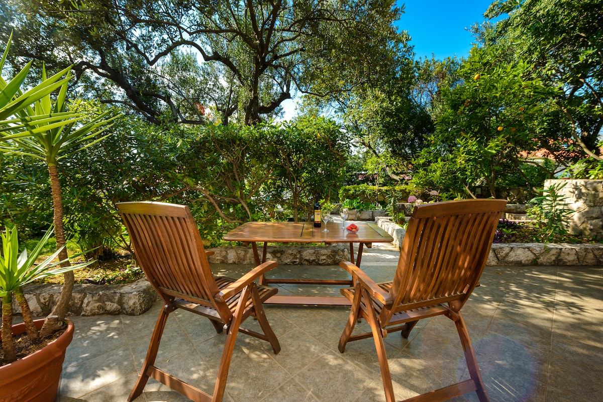 A serene outdoor area features wooden chairs positioned around a small table. Lush greenery surrounds the space, providing a tranquil setting. Sunlight filters through the leaves, creating a warm ambience. Decorative potted plants add a touch of nature to the peaceful retreat.