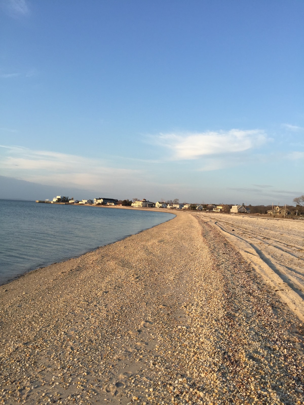 A sandy beach stretches along the shoreline, with gentle waves lapping at the water's edge. Soft clouds drift across a clear blue sky, creating a serene atmosphere. Beachfront homes are visible in the distance, framing the picturesque landscape.