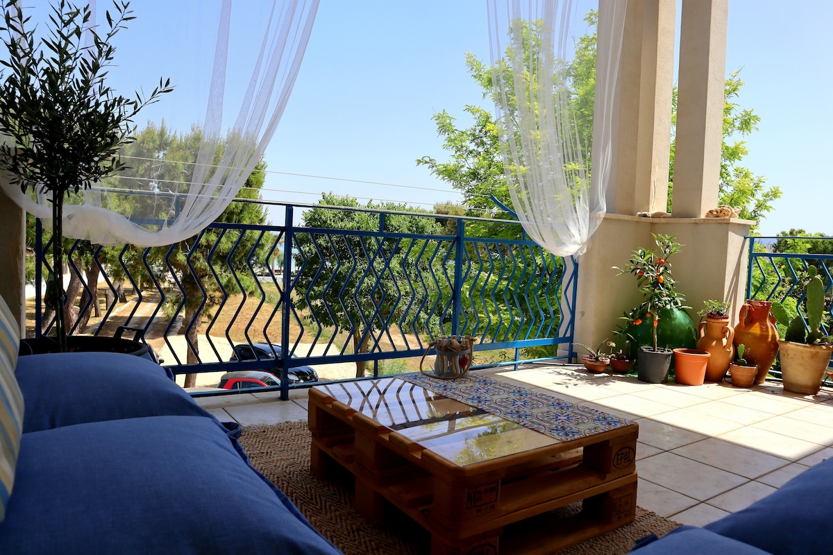A spacious private balcony features comfortable blue seating and a wooden coffee table adorned with mosaic details. Potted plants add greenery to the area, while light white curtains billow gently, offering a glimpse of the outdoor scenery.