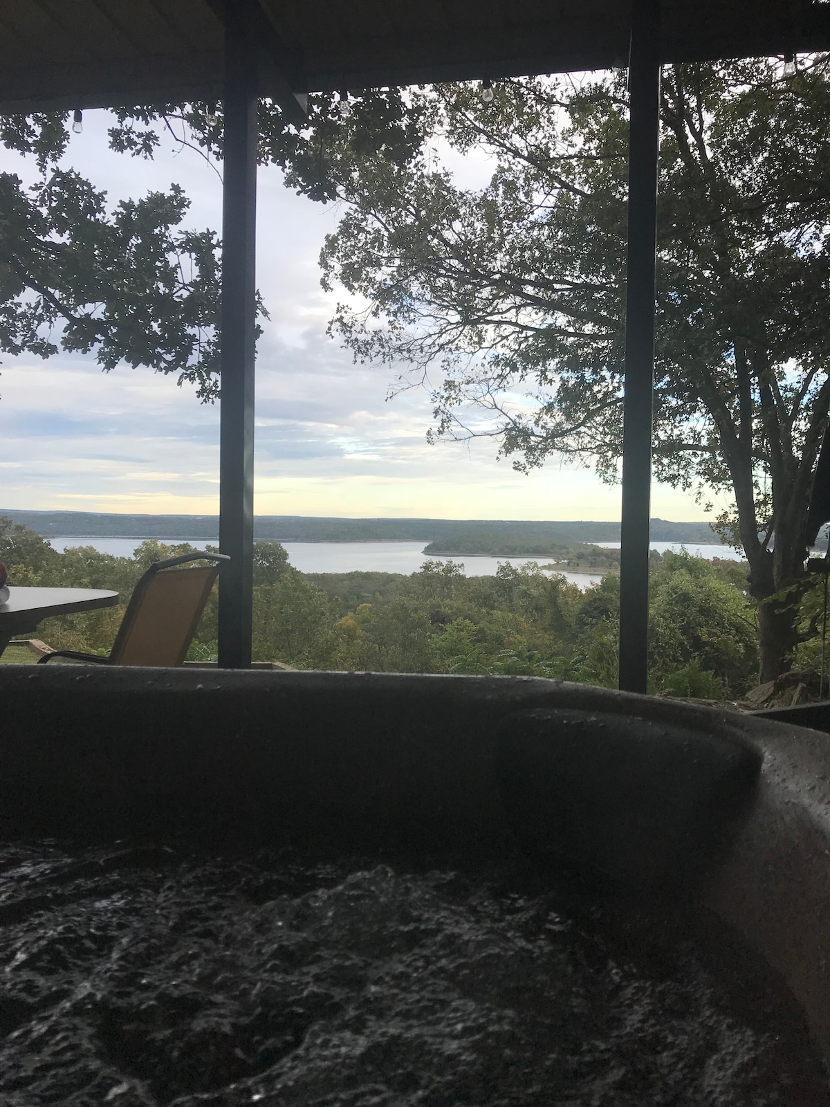 A view from a hot tub overlooks a serene landscape, with expansive water and distant hills visible in the background. Large windows create an open atmosphere, while the surrounding greenery adds to the natural setting.