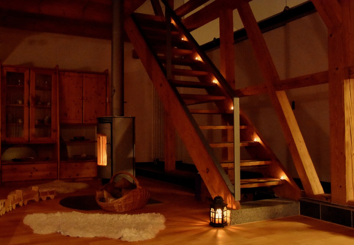 A warm and inviting interior features a wooden staircase illuminated by soft lighting. A stone fireplace stands nearby, accompanied by a woven basket and a lantern on the floor. Natural wood accents are visible throughout, enhancing the open and rustic environment.