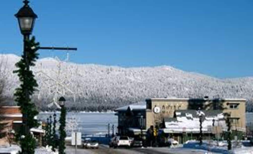 A winter scene captures a quaint town with snow-covered buildings. A mountain in the background is blanketed in white, contrasting against a clear blue sky. Streetlights and holiday decor line the road, hinting at a festive atmosphere. A frozen lake sits nearby.