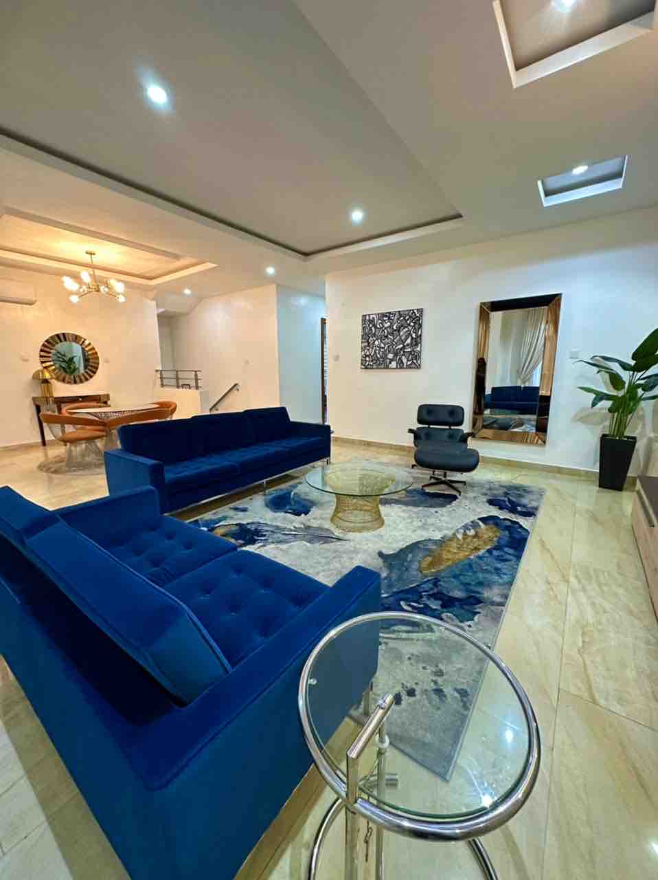 A stylish living area features two blue sofas facing a central glass coffee table. A large area rug adds texture to the floor, while a decorative mirror and a wall art piece enhance the ambiance. Natural light streams through a nearby window.