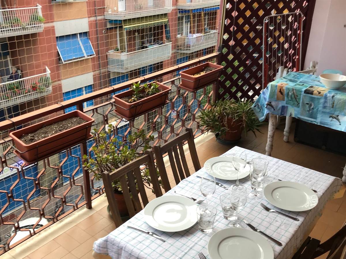 A dining table is set with plates and glasses, positioned on a terrace. Flower pots are lined along the protective railing, while a colorful tablecloth adds a touch of vibrancy. The neighboring buildings are visible in the background, showcasing a lively urban atmosphere.