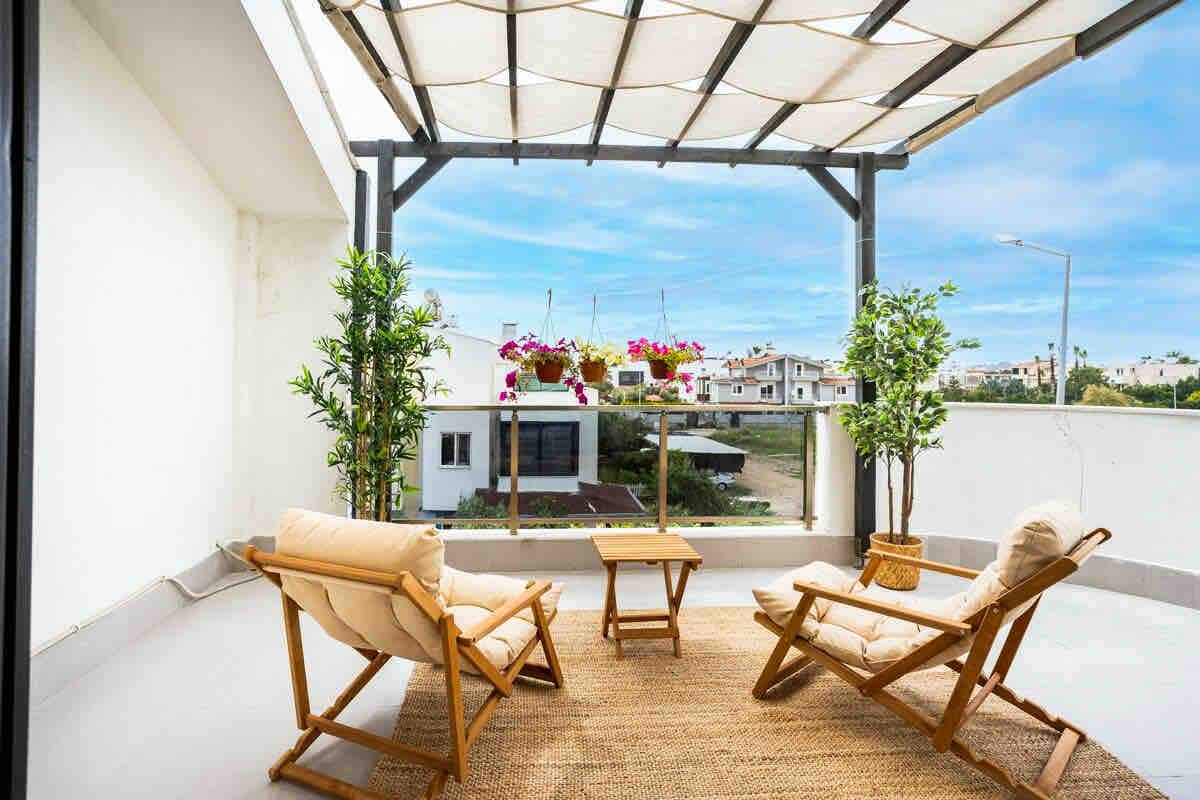 A spacious terrace is fitted with two wooden lounge chairs and a small table, surrounded by vibrant potted plants. The overhead pergola provides shade, while the open space offers views of the surrounding area under a bright blue sky.