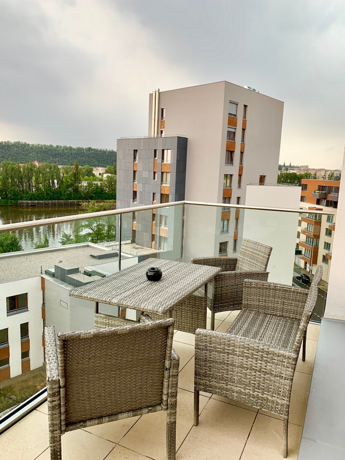 A balcony is furnished with a small table and two woven chairs, offering a view of the surrounding buildings and greenery. Glass railings provide safety while allowing unobstructed views. The floor is finished in light tiles, complementing the modern design of the area.