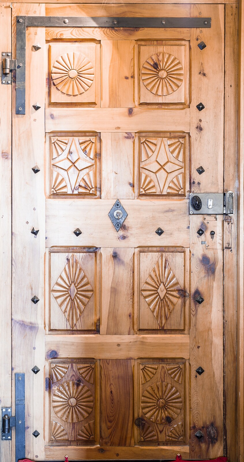 A traditional wooden door with intricate carvings and decorative elements is displayed. The door features a mix of geometric patterns, with metal accents and a rustic finish, providing an inviting entry point to the chalet.