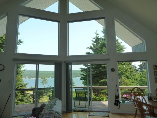 Large floor-to-ceiling windows create an open view of St. Ann's Bay, allowing natural light to fill the living space. The scenic landscape features lush trees and a deck with seating, offering a peaceful connection to the outdoors.