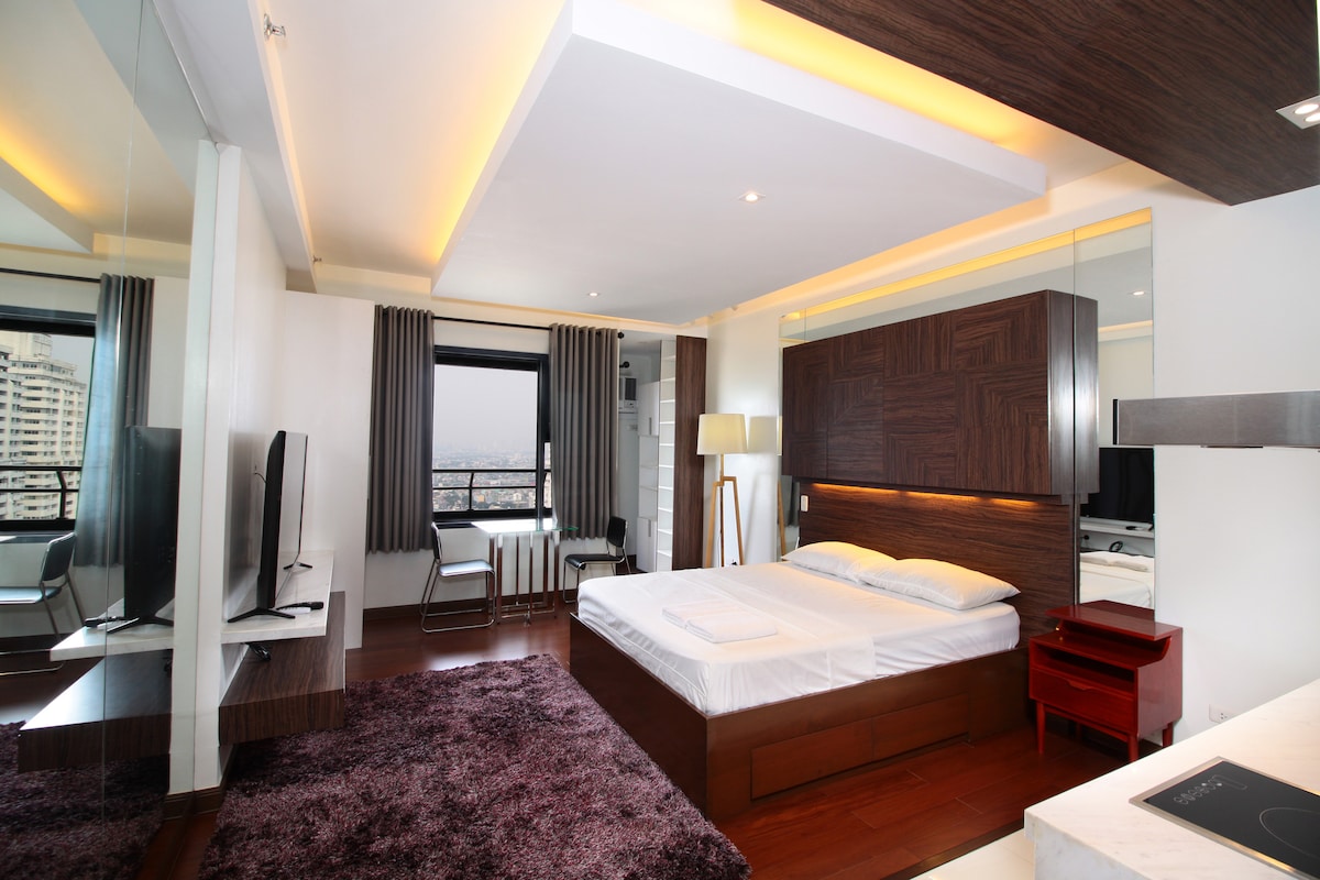 A studio apartment features a spacious bed against a wooden wall, with soft lighting enhancing the ambiance. A large window offers a view of the city skyline, while a TV is mounted on the opposite wall. A small dining table is located near the window.