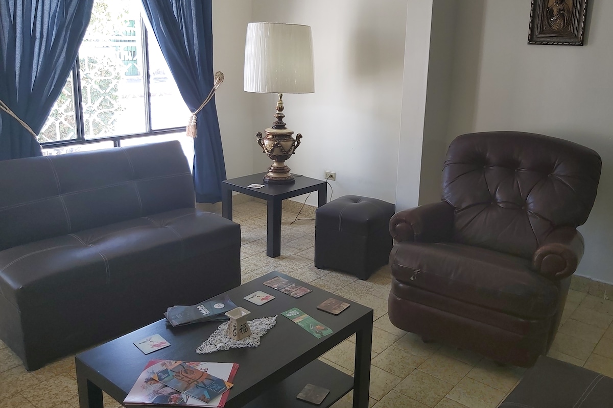 A living area features a black coffee table surrounded by a brown armchair and a sleek sofa. A decorative lamp stands beside the window, which is draped with blue curtains. Natural light filters into the space, highlighting the minimalistic furniture arrangement.