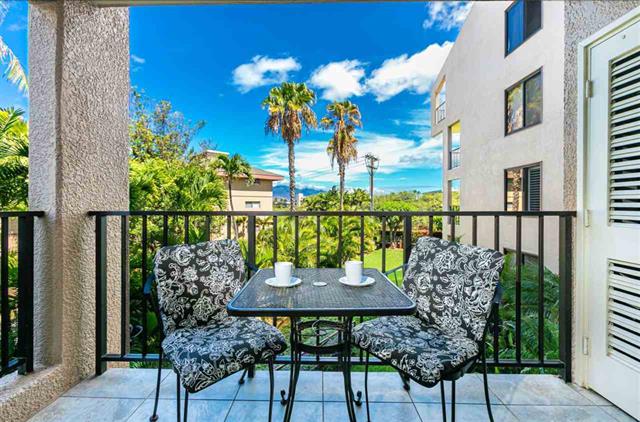 A small outdoor seating area is featured on the spacious lanai, furnished with a round table and two patterned chairs. Lush gardens and palm trees are visible in the background, complemented by a clear blue sky.