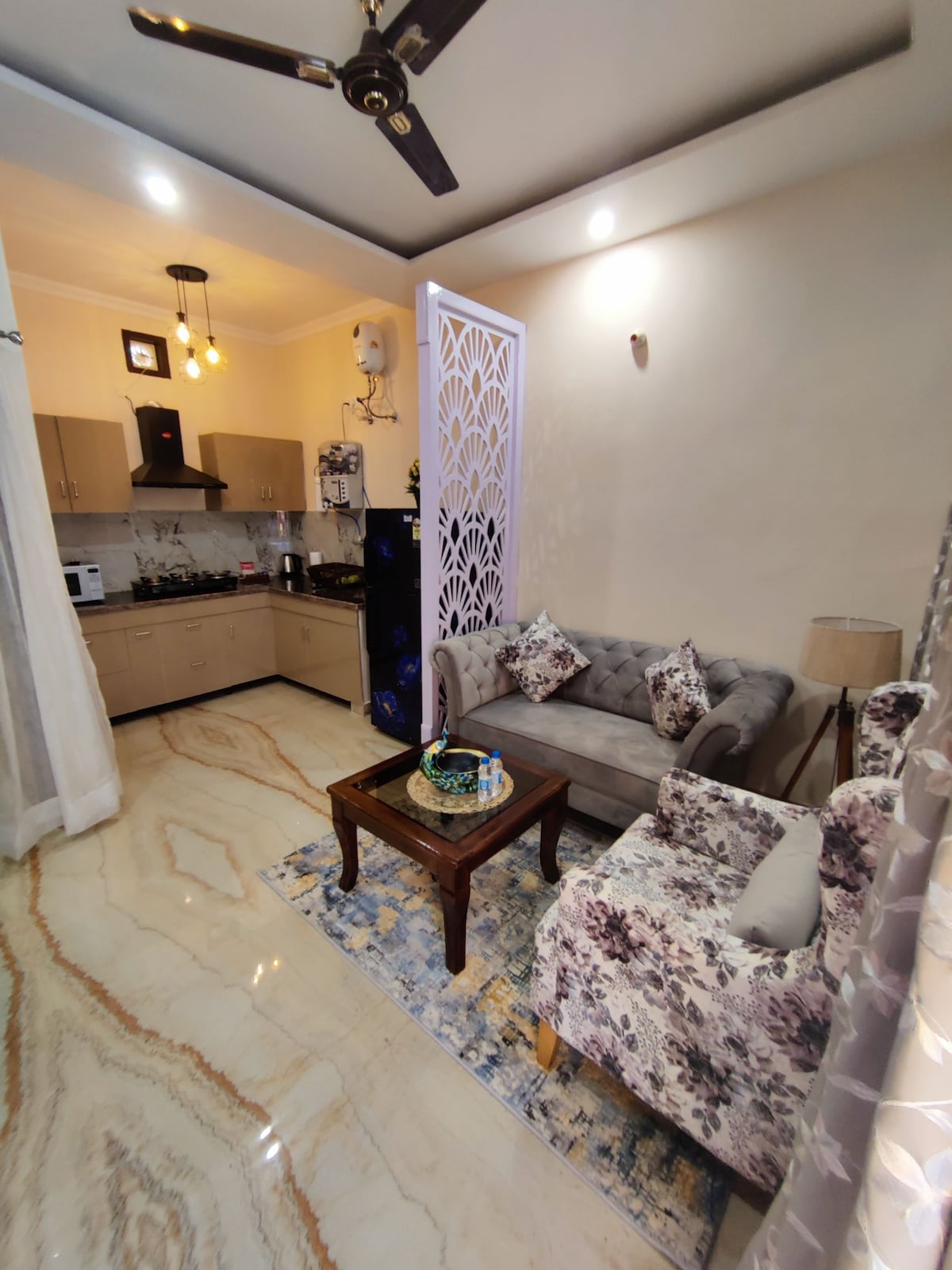 A cozy living area is presented with a plush grey sofa and a floral armchair, arranged around a wooden coffee table. A stylish rug adds texture to the polished floor. The modular kitchen is in view, featuring modern cabinets and appliances, with soft overhead lighting enhancing the warmth of the space.